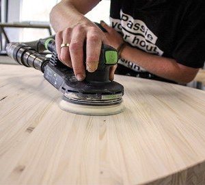 Super-Seat | Page Title | A person is sanding a round, light oak table with a black and green sander. |. | Hospitality Furniture