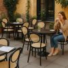 Super-Seat | Eine Frau sitzt an einem Cafétisch im Freien | Zwarte Thonet terrassenstoelen van gebogen hout en metaal, ronde tafels met glazen vaasjes, geel decor. | Horecameubilair