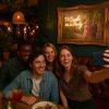 Super-Seat | Vier Freunde sitzen in einer gemütlichen Restaurantnische und lächeln für ein Selfie, während Getränke und Essen den Tisch füllen. | Donkerhouten-Tisch, Stoffbezug aus Stoff in warmem Taupe; Antikes Gold mit Neonkunst an der Wand. |. | Horecameubilair