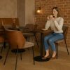 Super-Seat | New York designer chairs wood velour Mocha | Women sits on wooden mocha velour chair at round table, bottom wall, dried flowers in vase. | Hospitality Furniture