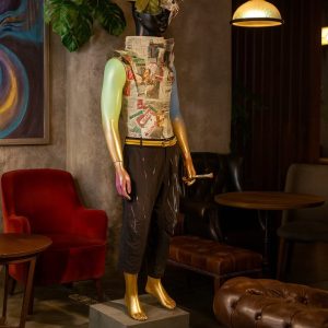 Super-Seat | Wastelander #4 | Zwartleren fauteuils, planten, duistere lounge; pop met gouden handen/voeten, zwart textiel, papieren hoed. | Horecameubilair